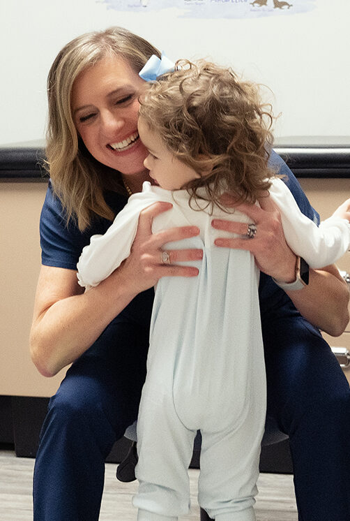 Dr. Beth Galla of Sweetbay Pediatrics facing the camera and smiling as she picks up a toddler with curly brown hair