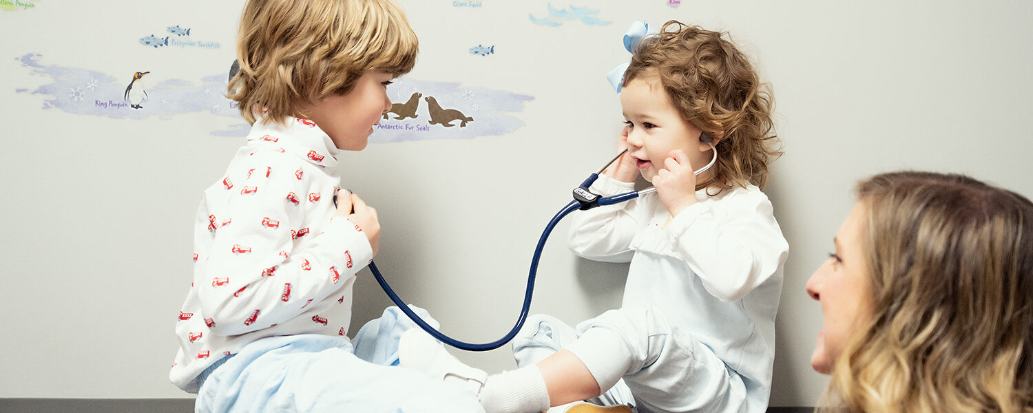 Two young caucasian patients of Dr. Beth Galla face each other sitting on the exam table as the younger child listens to their brother's heart through a stethoscope. Dr. Beth Galla looks at them, smiling.