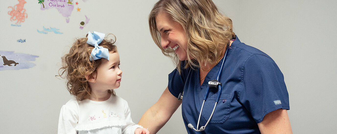 Dr. Beth Galla of Sweetbay Pediatrics, wearing blue scrubs, looking down and smiling at a toddler with curly brown hair 