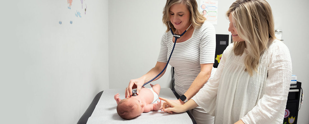 Dr. Beth Galla of Sweetbay Pediatrics places her stethoscope on the chest of a caucasian infant laying on the exam table while the infants mother, standing next to Dr. Galla, looks down at the child.